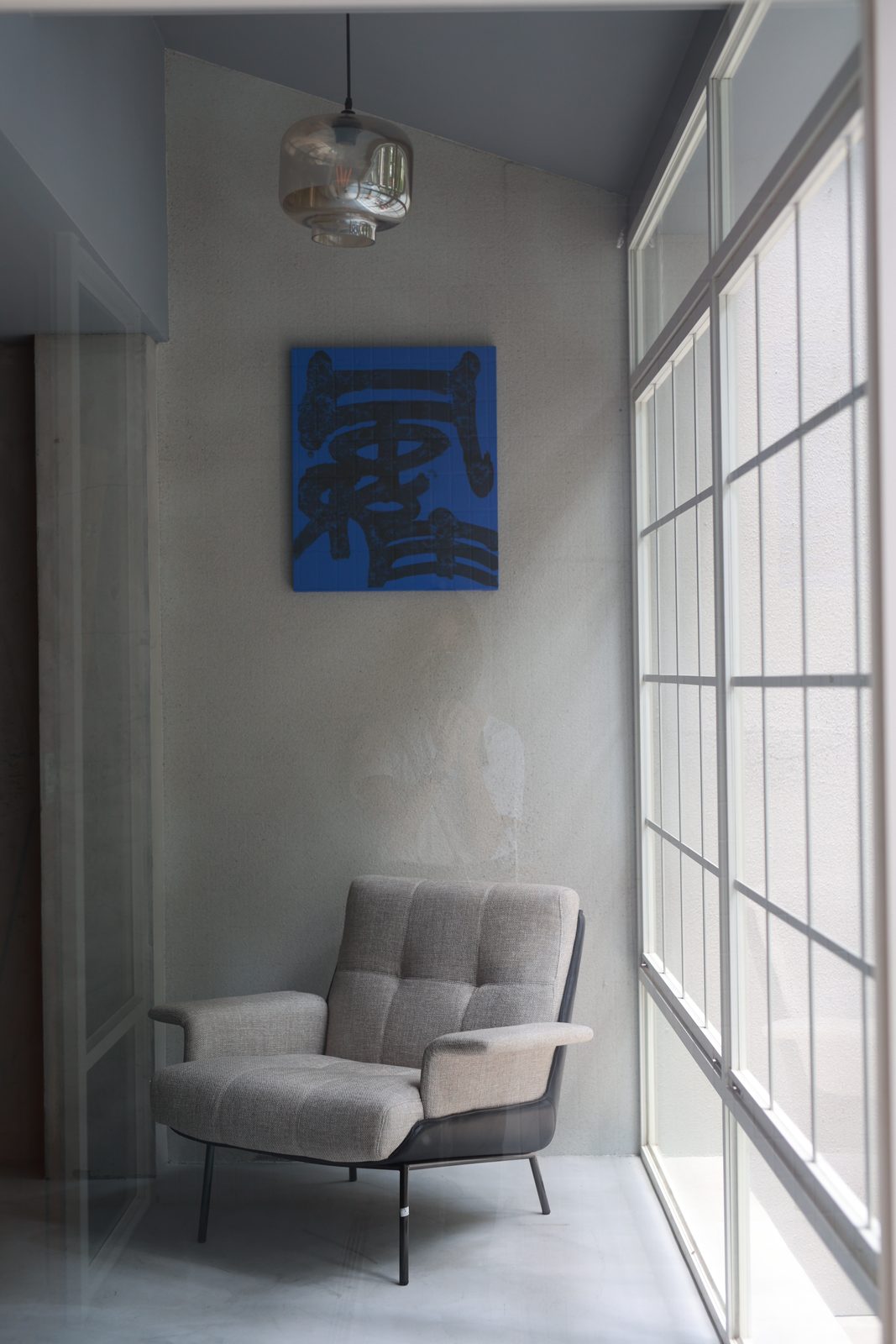 A blue-ground calligraphy on a concrete wall beside a tall steel window and a grey armchair
