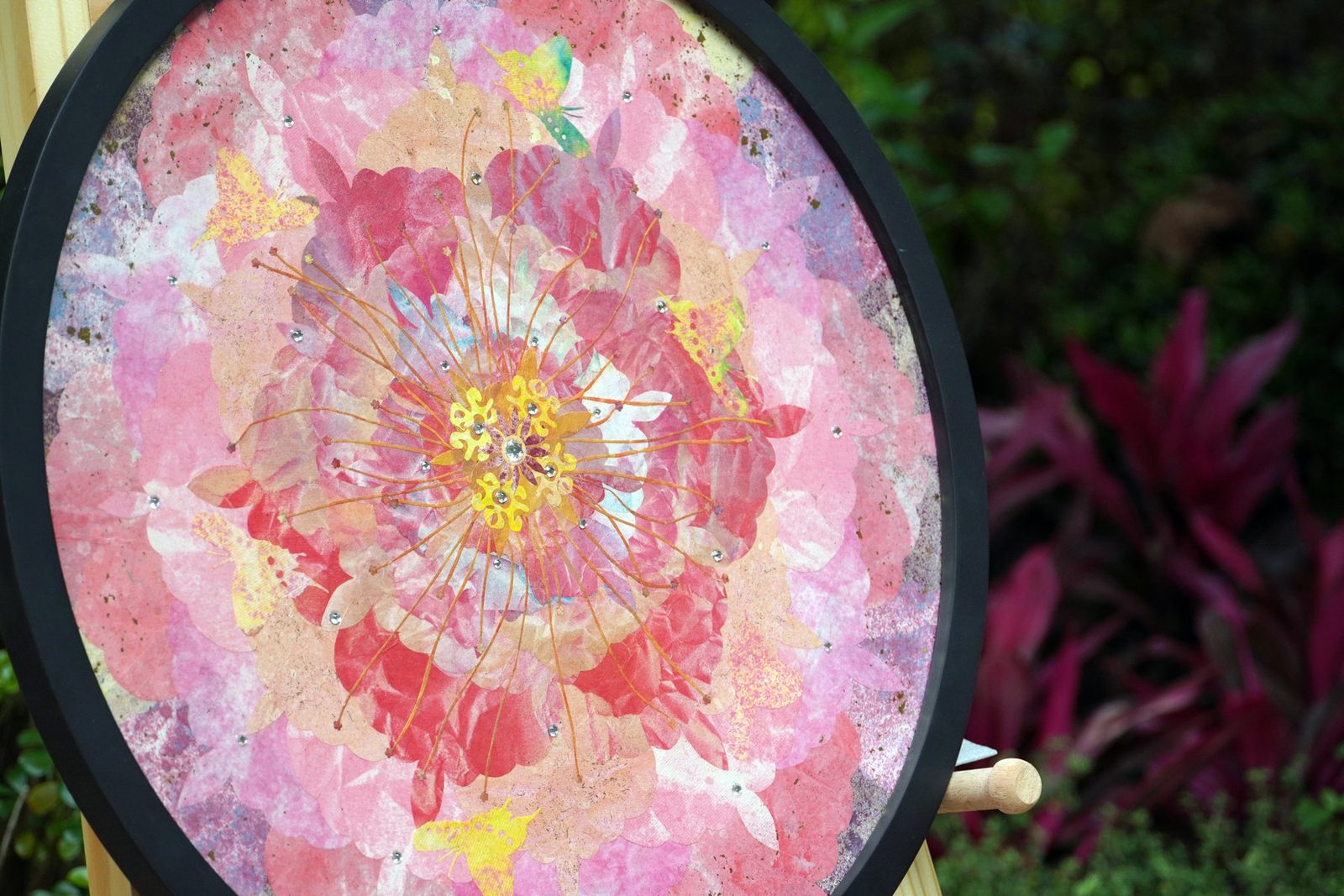 A round mineral painting in coral and rose, framed in black, among palms and ferns in a glass garden pavilion
