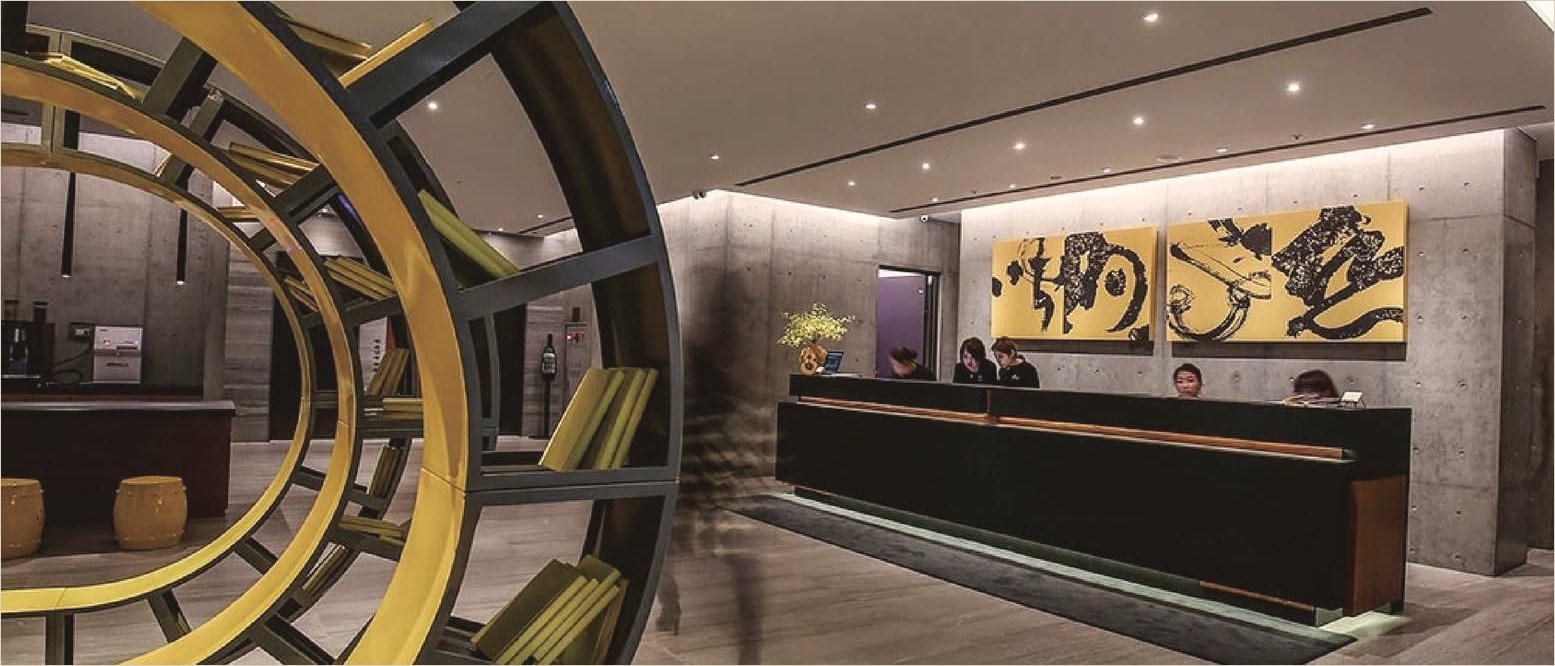 The same lobby by day: the yellow ring bookshelf at left, the long reception desk and the two gold-ground ink panels on the concrete wall