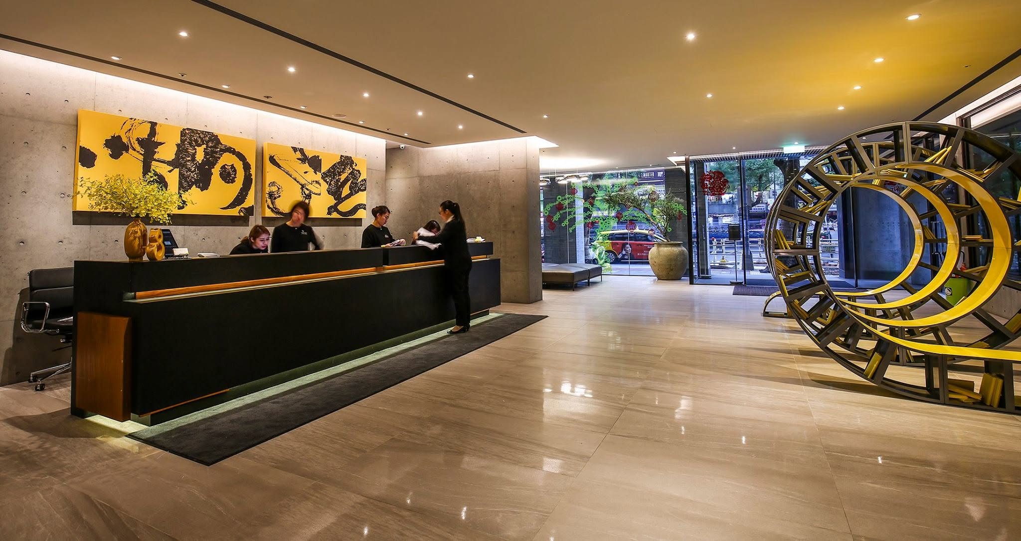 The lobby of CityInn Hotel on Fuxing North Road, Taipei, by day: two gold-ground panels of black ink calligraphy above the long reception desk on a concrete wall, a yellow ring-shaped bookshelf at right