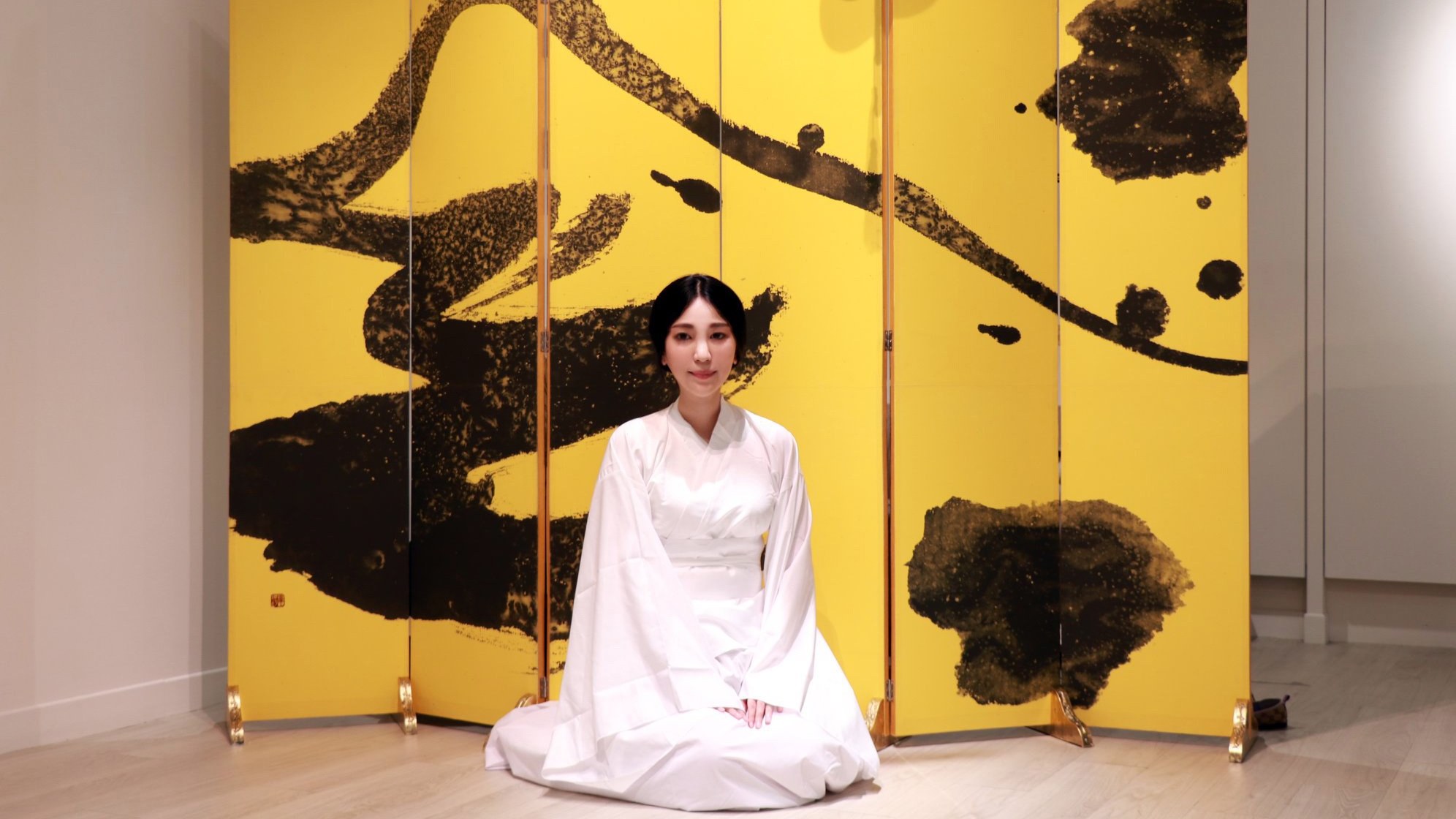 Ku Chi-Chun seated in a white robe before her gold six-panel screen painted in black ink