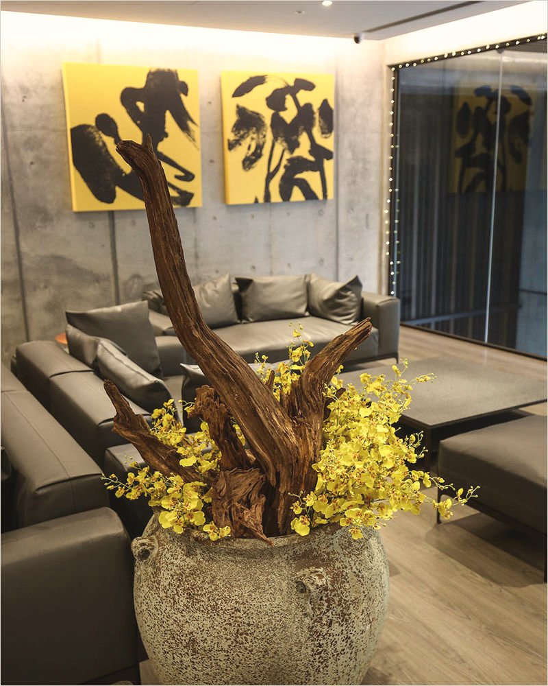 Hotel lounge: two square yellow canvases with black ink strokes above grey leather sofas, and a driftwood-and-orchid arrangement in a stone urn