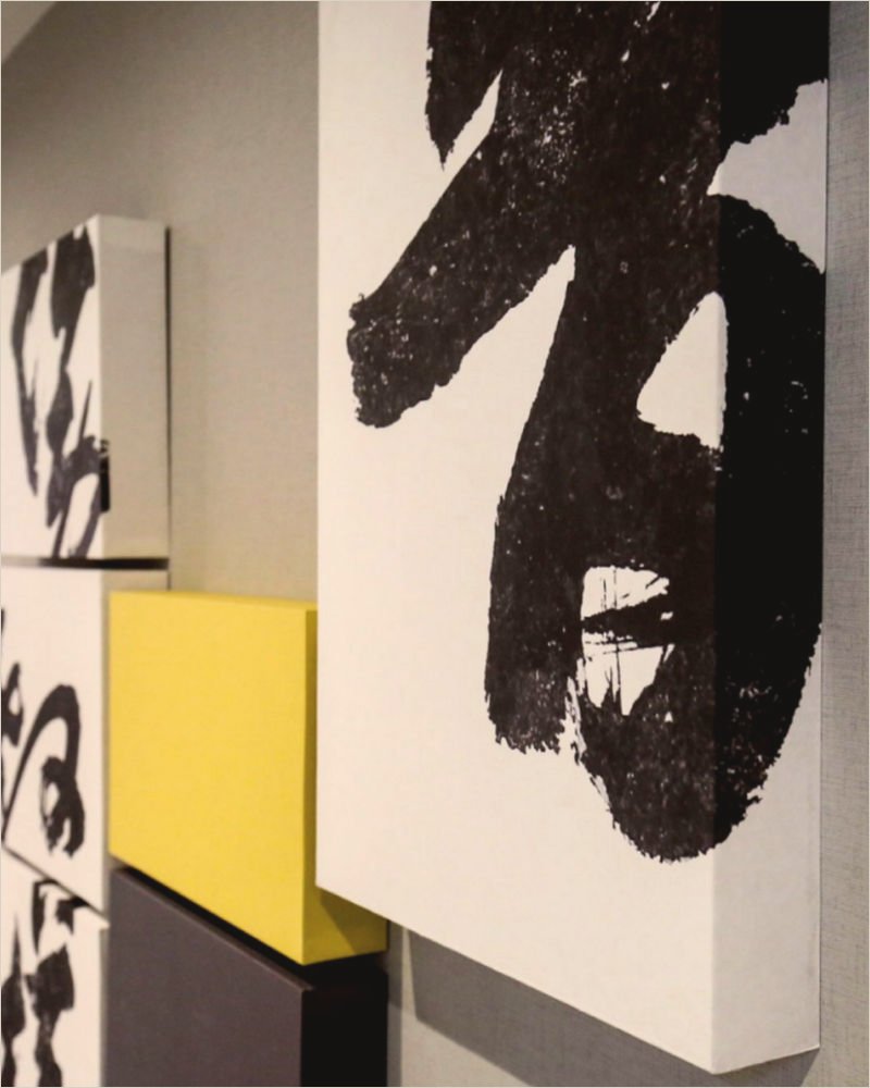 Close view of the corridor canvases: a single black ink character on white, a yellow block and a grey block stepping down the wall