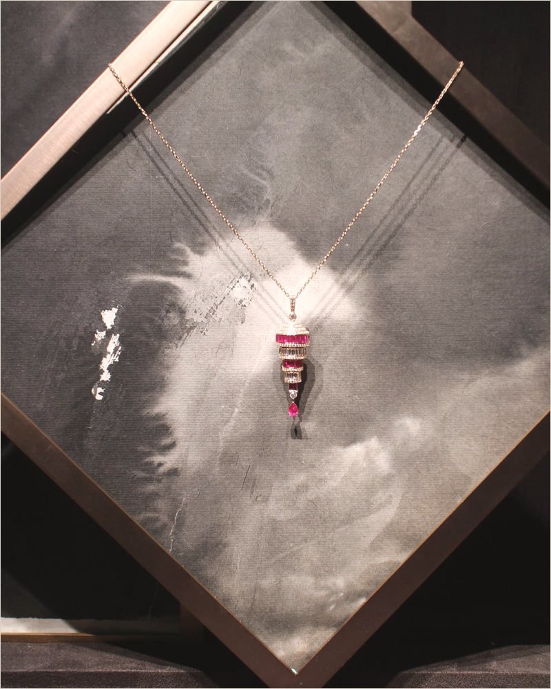 A ruby-and-diamond pendant on a gold chain hung over a square black-and-white ink-wash panel in a bronze frame, inside a lit display niche