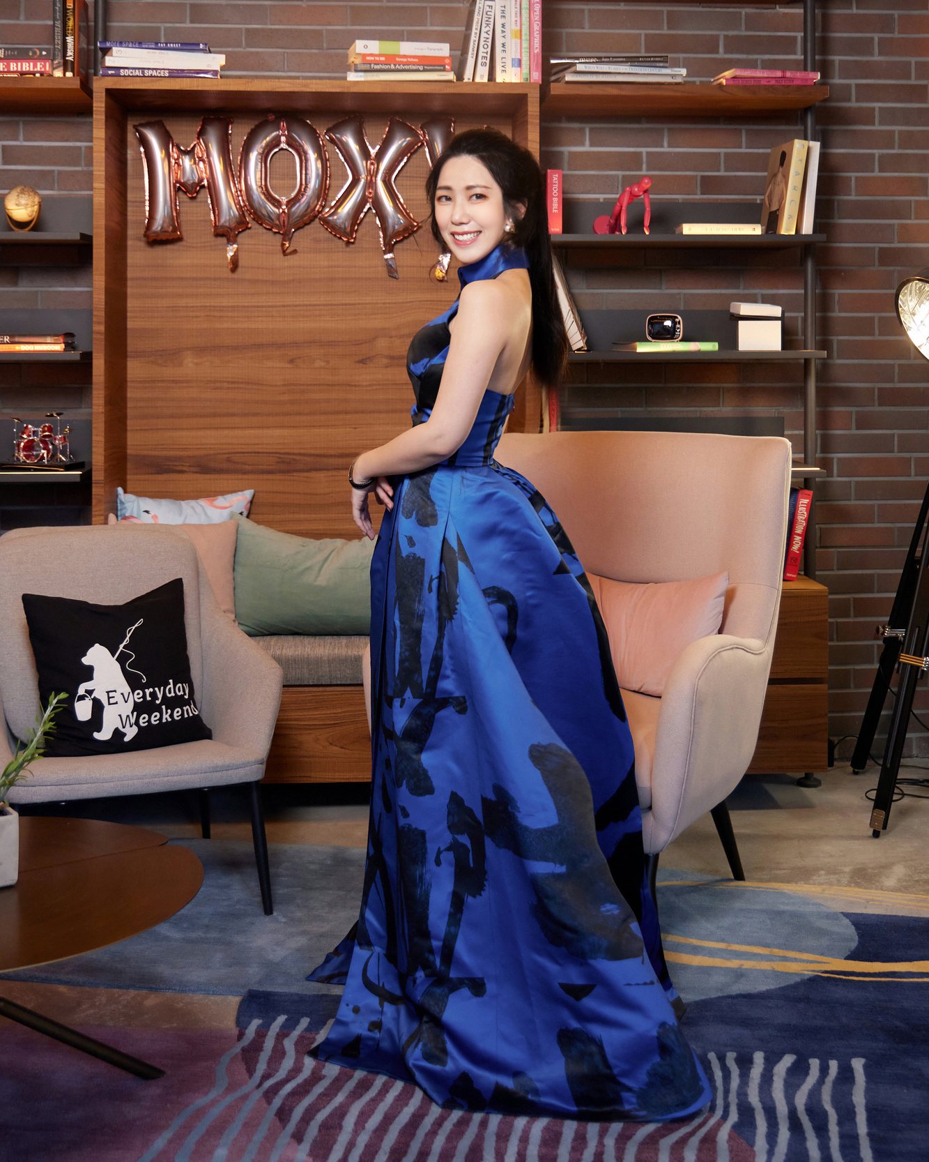 Ku Chi-Chun in a long sapphire-blue gown, looking back over her shoulder in a lounge with bookshelves