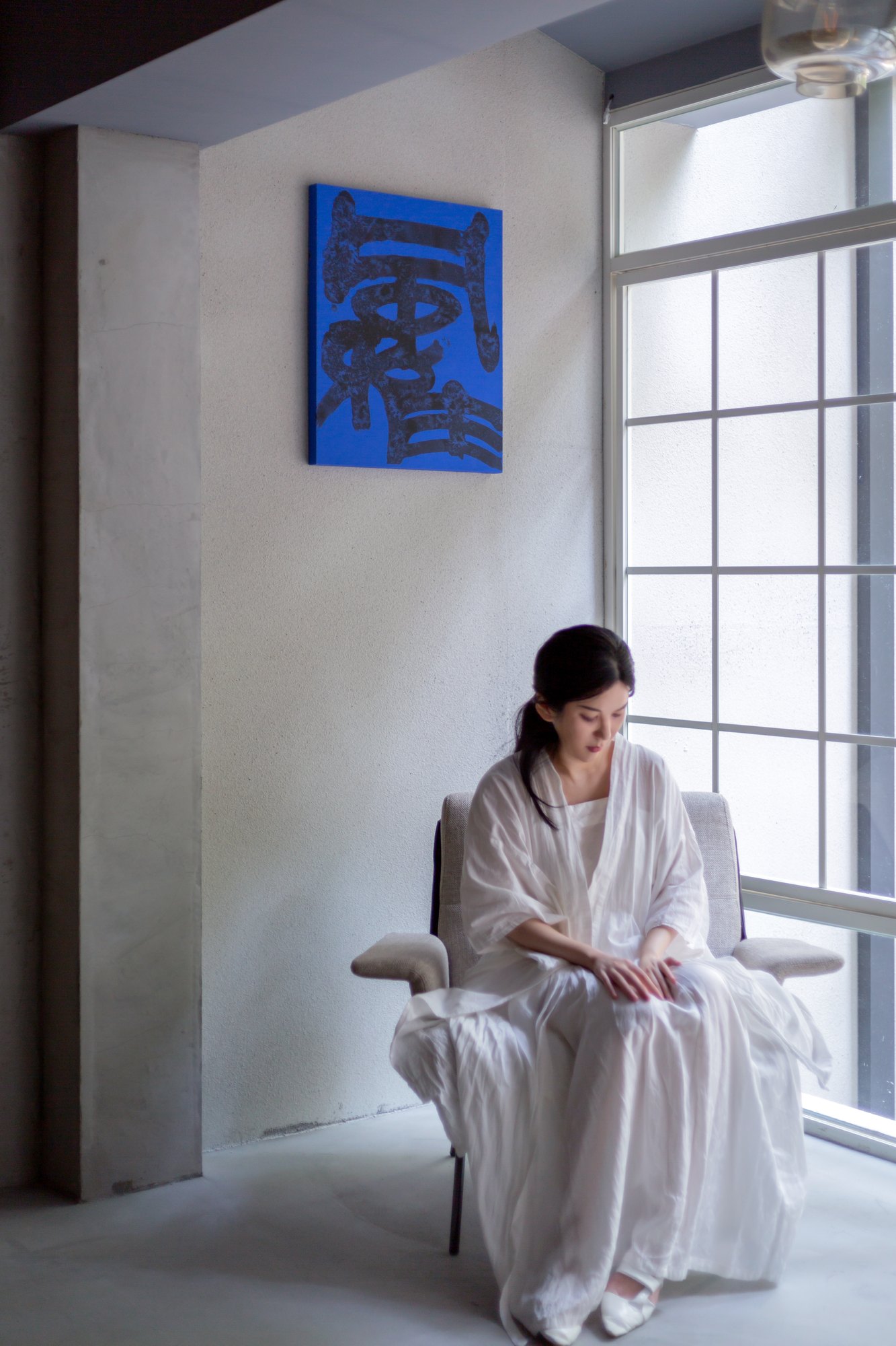 Ku Chi-Chun seated by a window in white, a blue calligraphy on the wall behind her