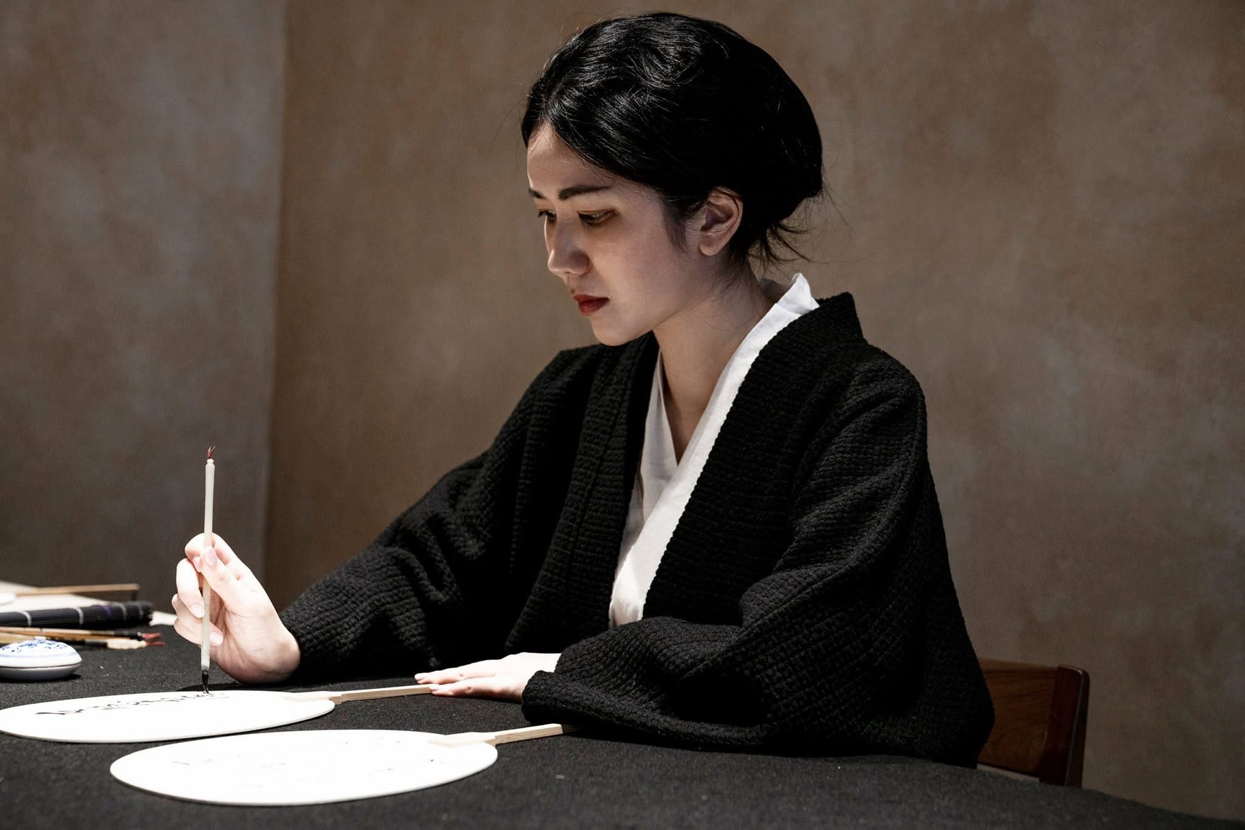 Ku Chi-Chun in a black jacket at a black-felt table, brushing calligraphy onto round white fans