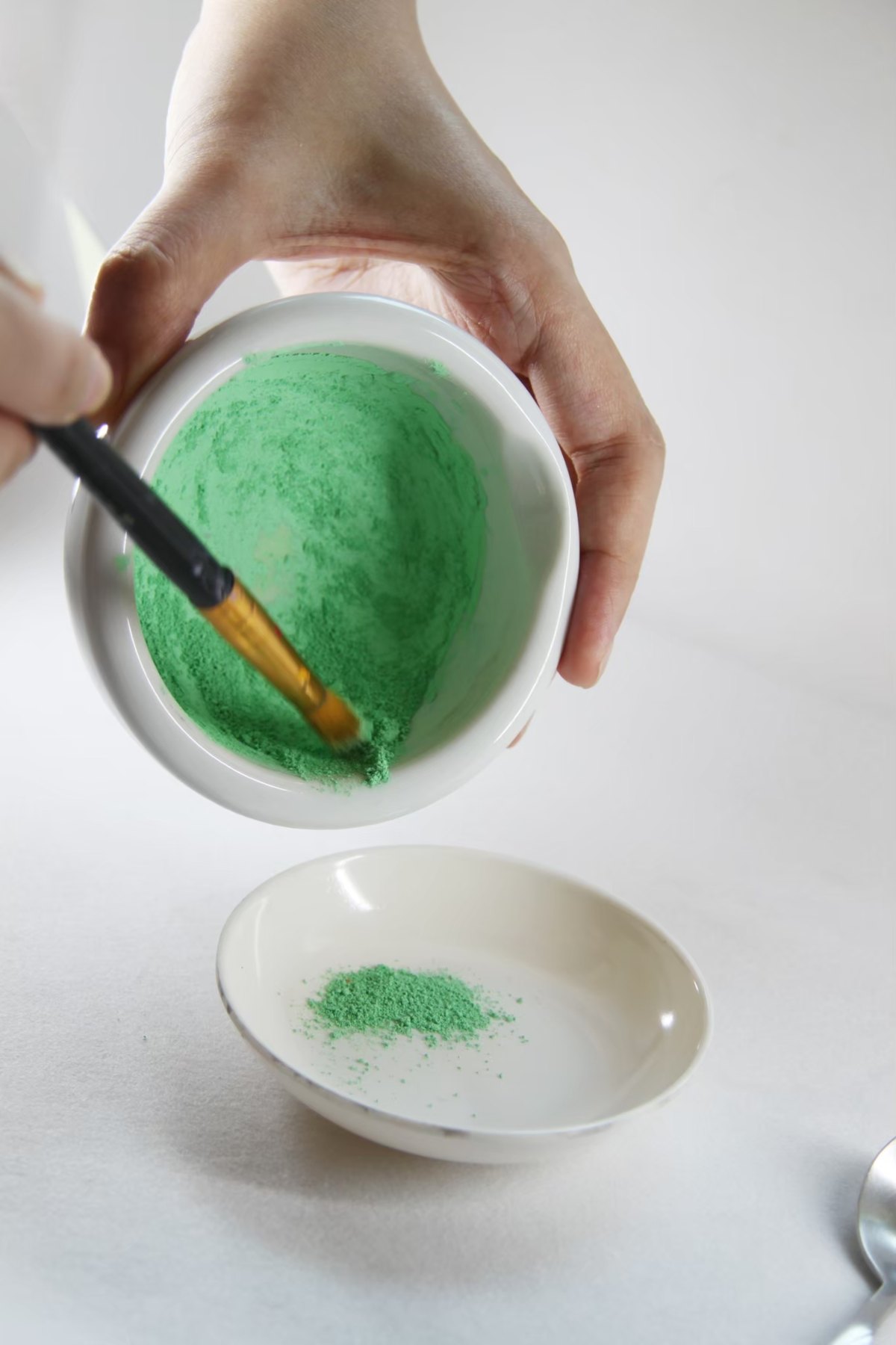 A hand working green pigment in a bowl with a brush