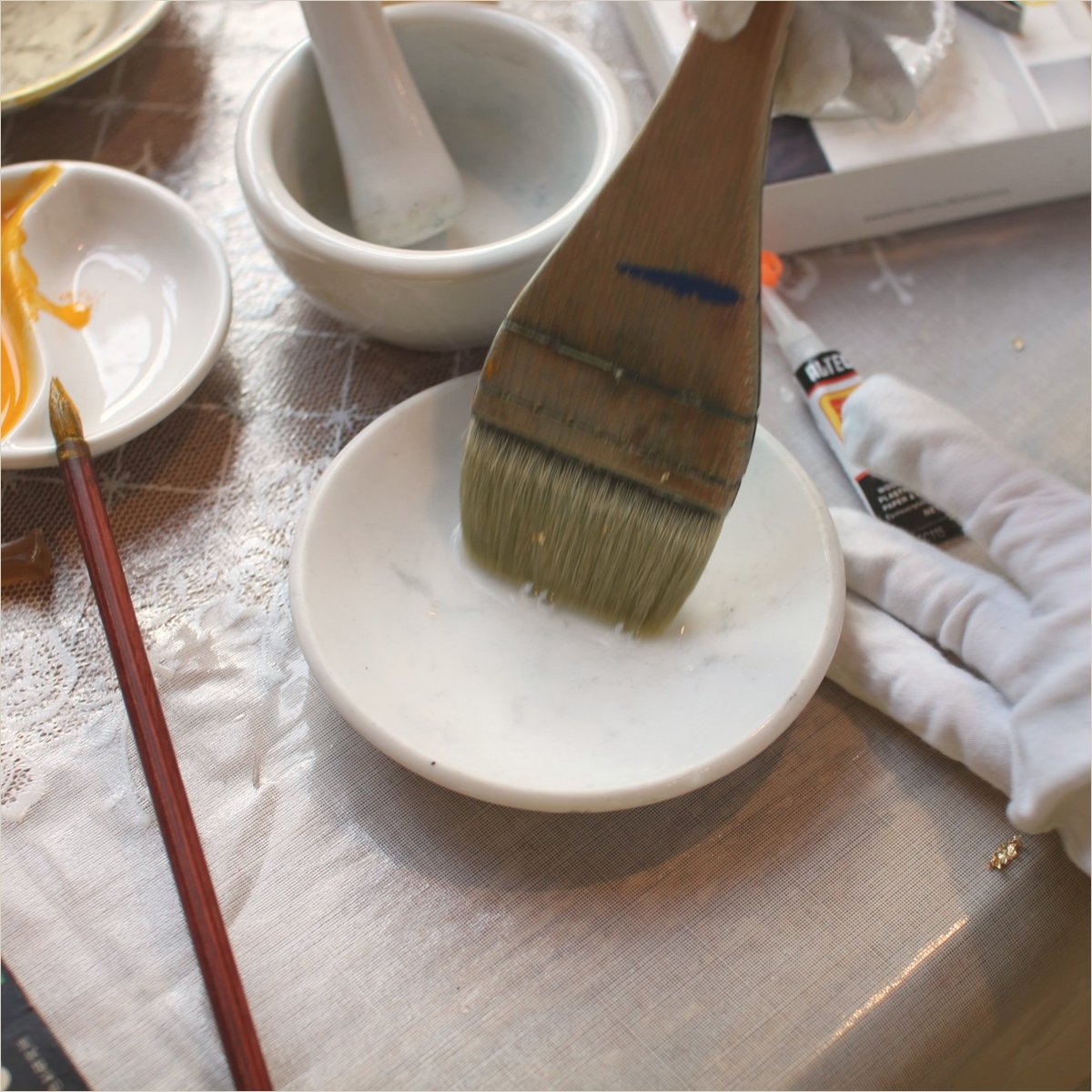 A wide brush, a dish of animal glue and a bowl of yellow pigment on lace