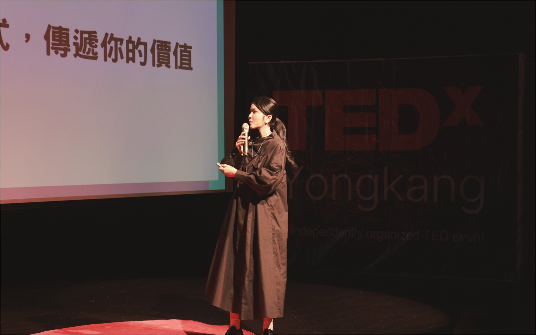 Ku Chi-Chun speaking into a microphone on the TEDxYongkang stage, the TEDx wordmark large behind her and the red circle at her feet