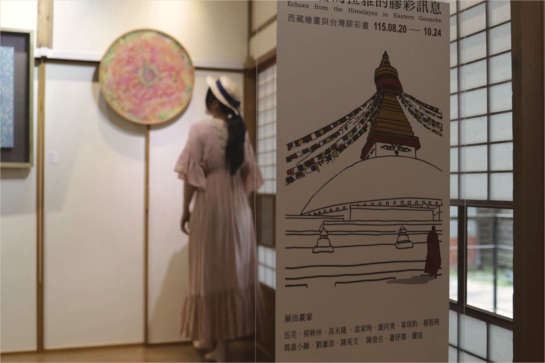 Ku Chi-Chun in a pink dress and straw hat, back to the camera, before her round mineral painting on the gallery wall, with the exhibition sign in the foreground