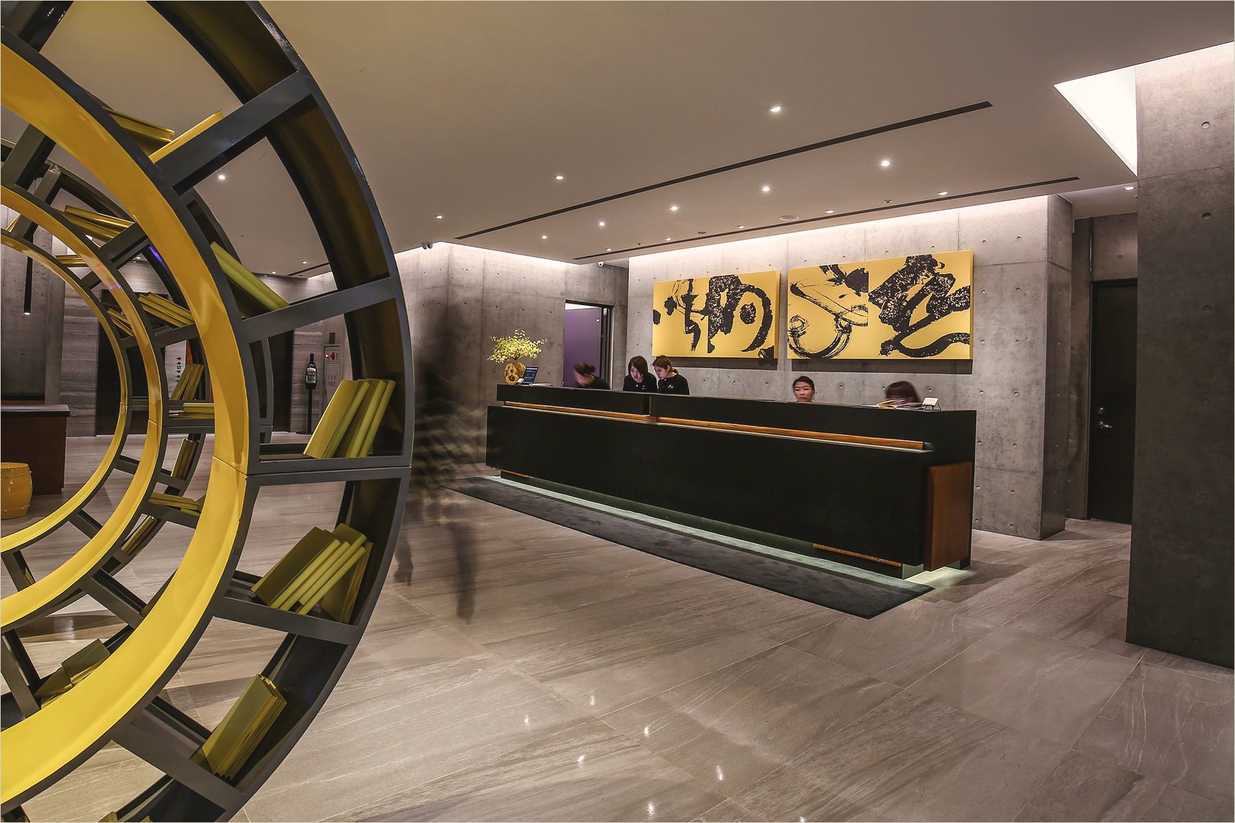 The lobby of CityInn Hotel on Fuxing North Road, Taipei, at night: two tall yellow panels of black ink calligraphy above the reception desk on a raw concrete wall, a yellow ring-shaped bookshelf in the foreground