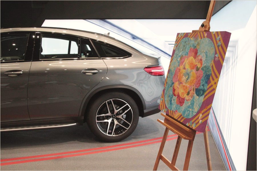 A mineral painting of coral and teal blossoms on a maroon ground, on a wooden easel beside the rear wheel of a grey Mercedes-Benz coupé in a showroom